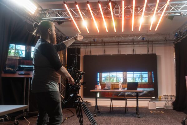 Joe Rees-Jones, creative technician stands in the XR Stories research and development lab, a studio space with specialist equipment including an LED wall, a video camera on a tripod and rows of LED lights on the ceiling.