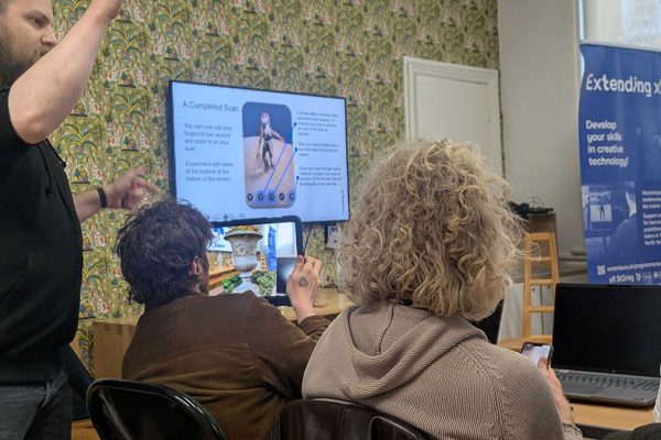 Teacher, Joe-Rees Jones stands next to participants at an Extending XR workshop looking towards a large TV screen and a roller banner.