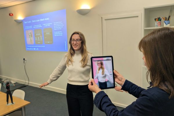 A person holding a tablet device takes a 3D scan of another person with long blonde hair standing with their arms outstretched at an Extending XR creative technology skills development workshop.
